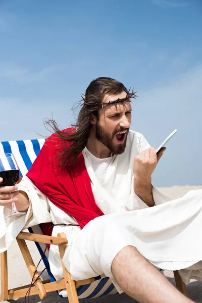 Angry Jesus sitting on sun lounger with glass of wine and screaming at smartphone in desert — Stock Photo