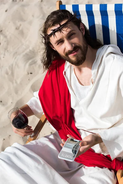 Jesus resting on sun lounger with glass of wine and holding smartphone with tickets website in desert — Stock Photo
