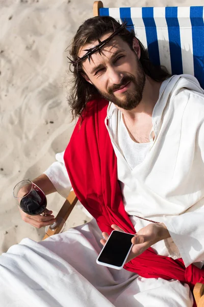 Jesus resting on sun lounger with glass of wine and holding smartphone with blank screen in desert — Stock Photo