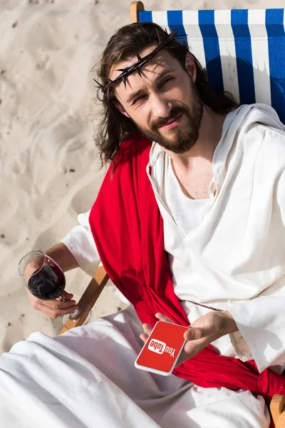 Jesus resting on sun lounger with glass of wine and holding smartphone with youtube page in desert — Stock Photo