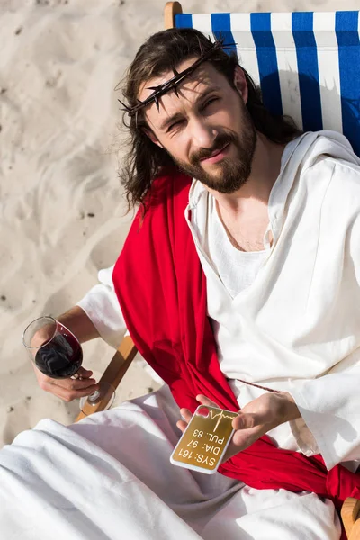Jesus resting on sun lounger with glass of wine and holding smartphone with medical appliance in desert — Stock Photo