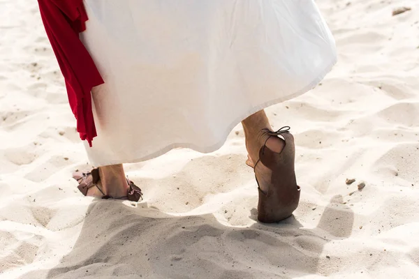 Обрезанный образ Иисуса в халате, сандалиях и красной ленте, идущих по песку в пустыне — стоковое фото