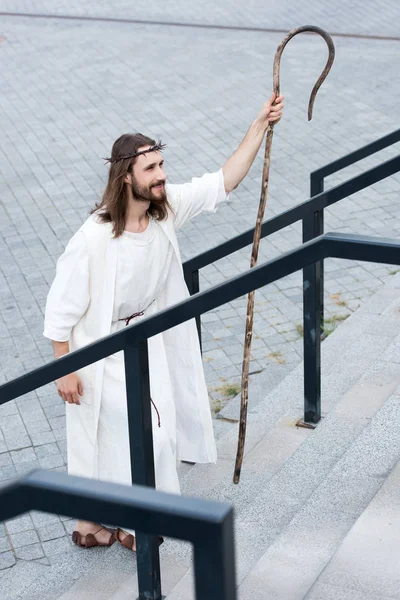 High angle view of cheerful Jesus in robe and crown of thorns walking on stairs with staff — Stock Photo