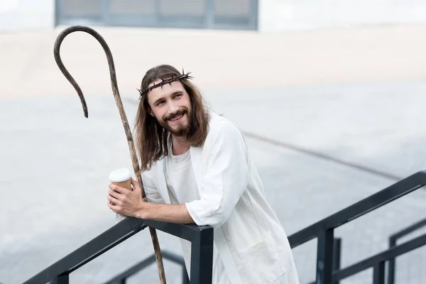 Smiling Jesus in robe and crown of thorns holding disposable coffee cup and leaning on railing on street — Stock Photo