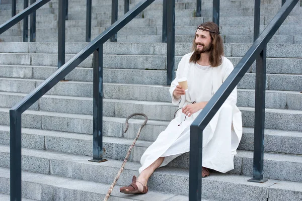 Cheerful Jesus in robe and crown of thorns sitting on stairs and holding disposable coffee cup on street — Stock Photo