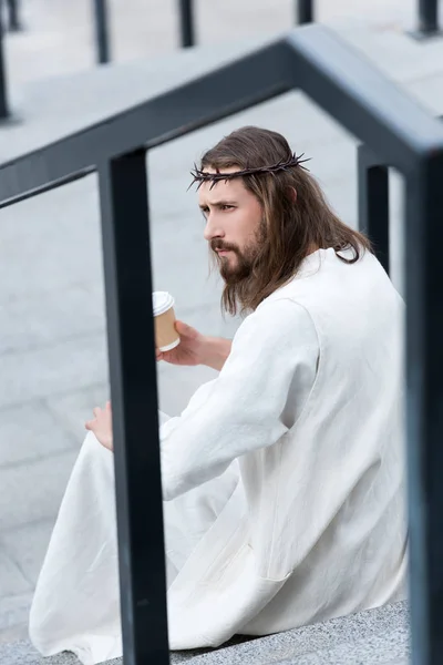 Боковой вид на серьезного Иисуса в мантии и терновом венце, сидящего на лестнице и держащего одноразовую чашку кофе на улице — стоковое фото