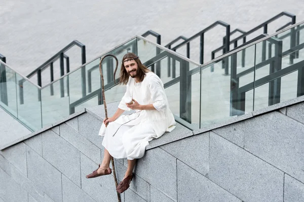 Улыбающийся Иисус в мантии и терновом венце, сидящий на лестнице и держащий кофе в бумажной чашке — стоковое фото