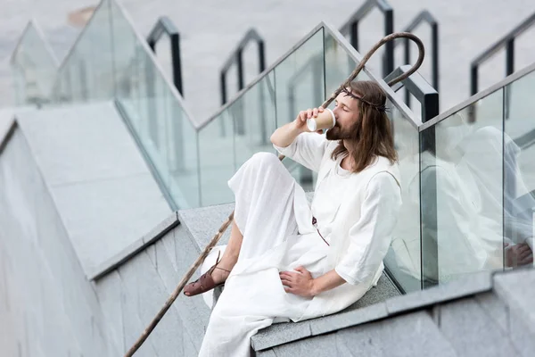 Jesus in robe and crown of thorns sitting on staircase side and drinking coffee from disposable coffee cup — Stock Photo