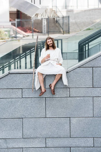Cheerful Jesus in robe and crown of thorns sitting on staircase side and holding coffee to go — Stock Photo