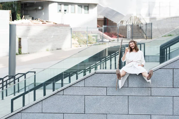 Щасливий Ісус в халаті і корона з терну, сидячи на сходах і показуючи чашку одноразової кави — стокове фото