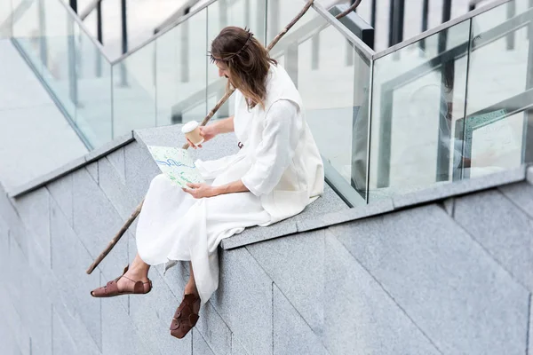Jesus in robe and crown of thorns sitting on staircase side, holding disposable coffee cup and looking at map — Stock Photo