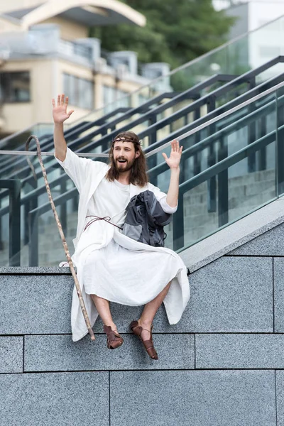 Happy Jesus in robe and crown of thorns sitting on staircase side and waving hands — Stock Photo