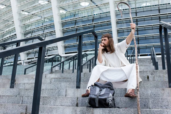 Pensive Jesus in robe and crown of thorns sitting with travel bag and staff on stairs of stadium — Stock Photo