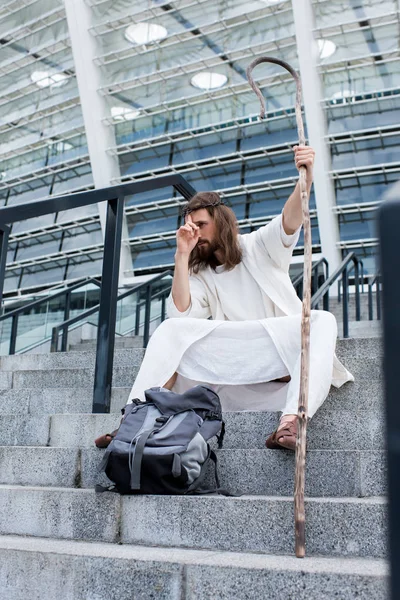 Jesus in robe sitting with travel bag and staff on stairs of stadium and fixing crown of thorns — Stock Photo