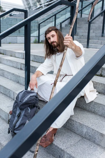 Jesus in Robe und Dornenkrone sitzt mit Reisetasche und Stab auf der Treppe und blickt in die Kamera — Stockfoto