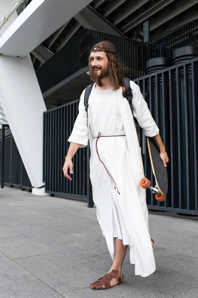 Smiling Jesus in robe, crown of thorns and bag holding skateboard on street — Stock Photo