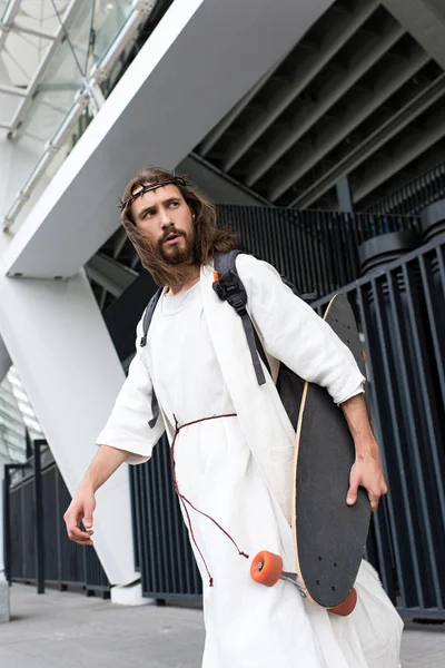 Low angle view of Jesus in robe, crown of thorns and bag walking with skateboard on street — Stock Photo