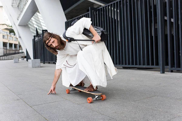 Jesus in robe and crown of thorns skating on longboard on street — Stock Photo