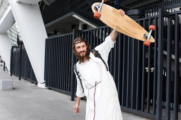 Cheerful Jesus in robe, crown of thorns and bag showing skateboard on street — Stock Photo