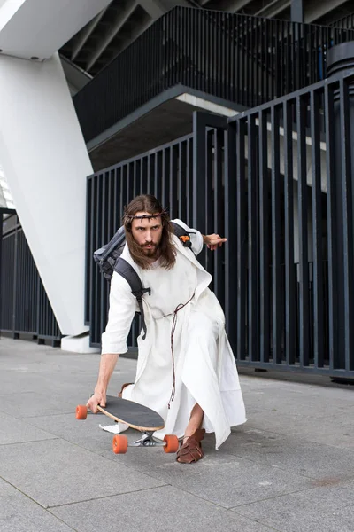 Серьезный Иисус в мантии и терновом венце катается на коньках на длинном борде по улице — стоковое фото
