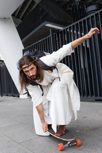 Handsome Jesus in robe and crown of thorns skating on longboard on street — Stock Photo