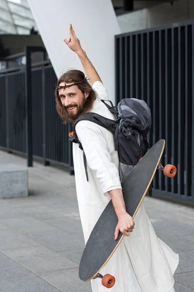 Smiling Jesus in robe and crown of thorns walking with longboard and showing two fingers up — Stock Photo
