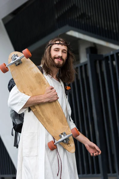 Low angle view of cheerful Jesus in robe and crown of thorns holding skateboard — Stock Photo