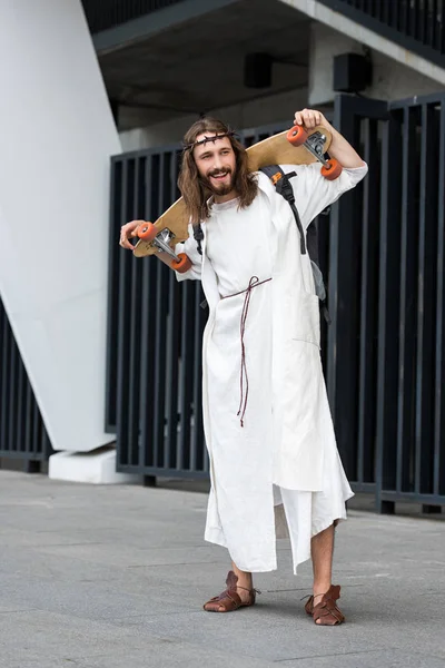 Smiling Jesus in robe and crown of thorns holding skateboard on shoulders — Stock Photo