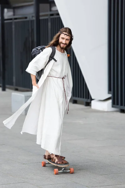 Smiling Jesus in robe and crown of thorns skating on longboard on street — Stock Photo