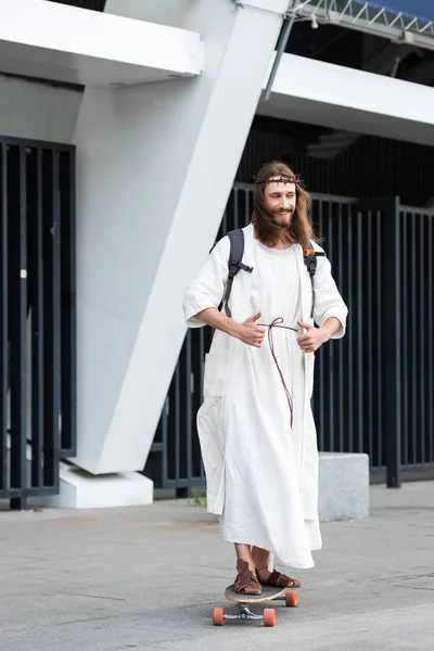 Улыбаясь, Иисус в халате и терновом венце катается на коньках на длинной доске по улице и показывает большие пальцы вверх — стоковое фото