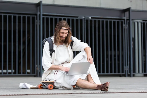Улыбающийся Иисус в мантии и терновом венце сидит на скейтборде и использует ноутбук в городе — стоковое фото