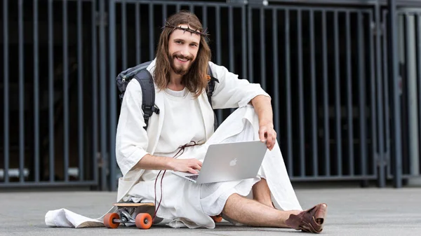 Cheerful Jesus in robe and crown of thorns sitting on skateboard and using laptop in city — Stock Photo
