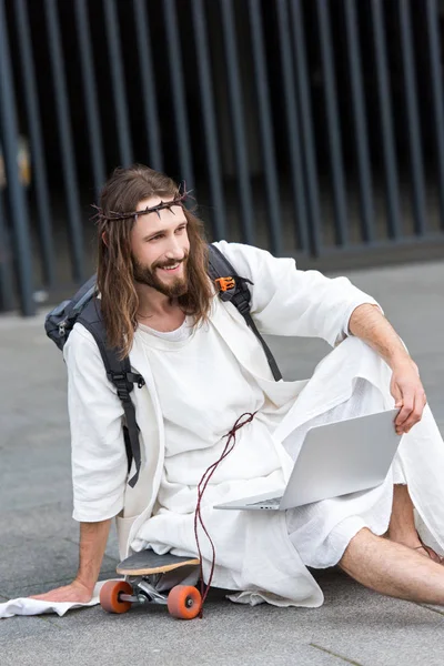 Smiling Jesus in robe and crown of thorns sitting on skateboard with laptop and looking away in city — Stock Photo