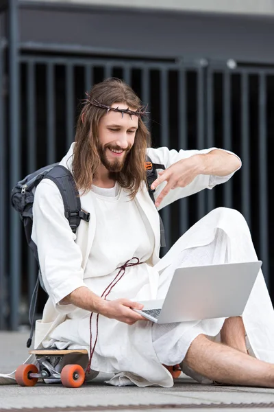 Jesus in robe and crown of thorns sitting on skateboard and showing two fingers during video call — Stock Photo