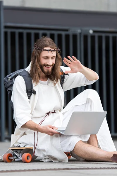 Smiling Jesus in robe and crown of thorns sitting on skateboard and waving hand during video call — Stock Photo