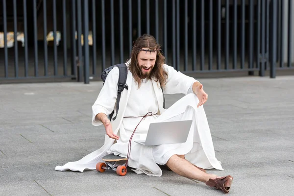 Irritated Jesus in robe and crown of thorns sitting on skateboard and gesturing to laptop in city — Stock Photo