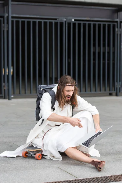 Angry Jesus in robe and crown of thorns sitting on skateboard and gesturing to laptop in city — Stock Photo