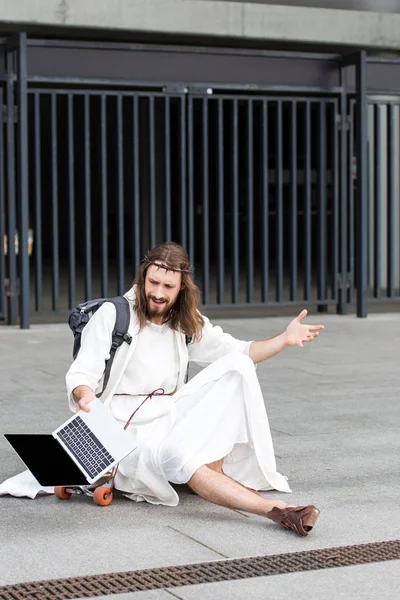 Раздраженный Иисус в мантии и терновом венце сидел на скейтборде и жестом к ноутбуку с чистым экраном в городе — стоковое фото