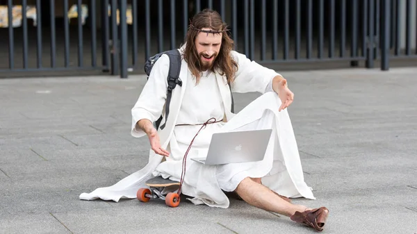 Irritated Jesus sitting on skateboard and gesturing to laptop on street — Stock Photo
