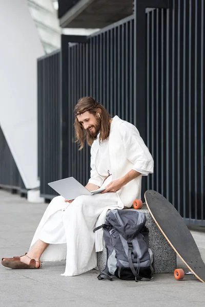 Cheerful Jesus in robe and crown of thorns sitting on stone and using laptop on street — Stock Photo