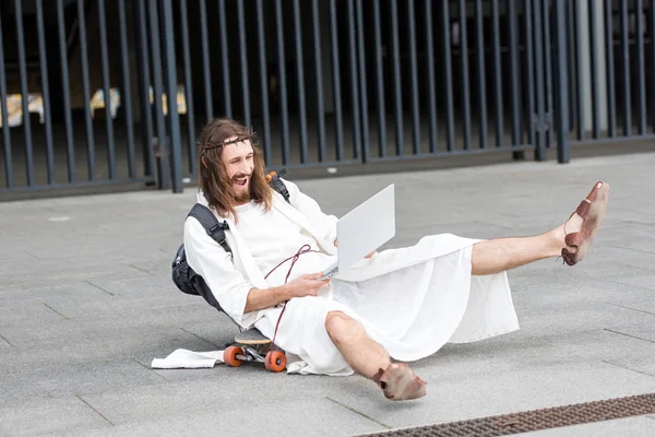 Laughing Jesus in robe and crown of thorns sitting on skateboard and using laptop in city — Stock Photo