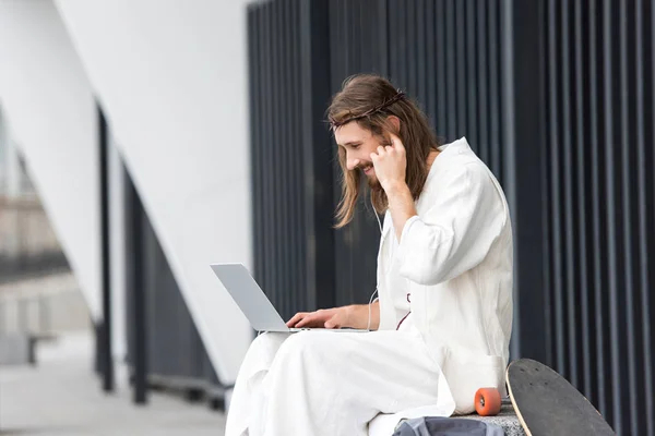 Side view of Jesus in robe and crown of thorns sitting on stone and using laptop on street — Stock Photo