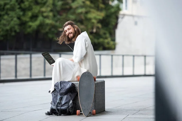 Jesus in robe and crown of thorns sitting on stone and holding laptop with html code on street — Stock Photo