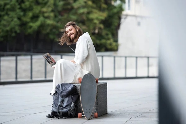 Jesus in robe and crown of thorns sitting on stone and using laptop with online booking website on street — Stock Photo