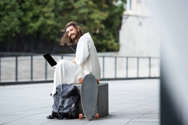 Smiling Jesus in robe and crown of thorns sitting on stone and using laptop with blank screen on street — Stock Photo