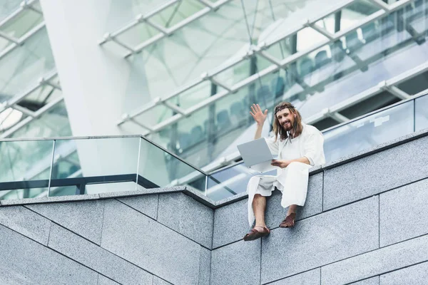 Low angle view of Jesus in robe and crown of thorns sitting on wall, using laptop and waving hand — Stock Photo