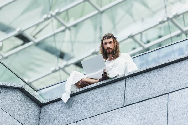 Vue à angle bas de Jésus sérieux en robe et couronne d'épines à l'aide d'un ordinateur portable assis sur le mur — Photo de stock