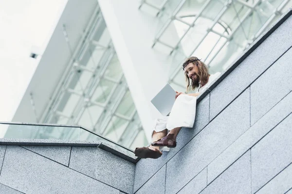 Vue à angle bas de Jésus souriant en robe et couronne d'épines à l'aide d'un ordinateur portable assis sur le mur — Photo de stock