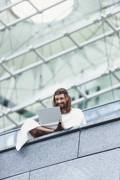Low angle view of laughing Jesus in robe and crown of thorns using laptop while sitting on wall — Stock Photo