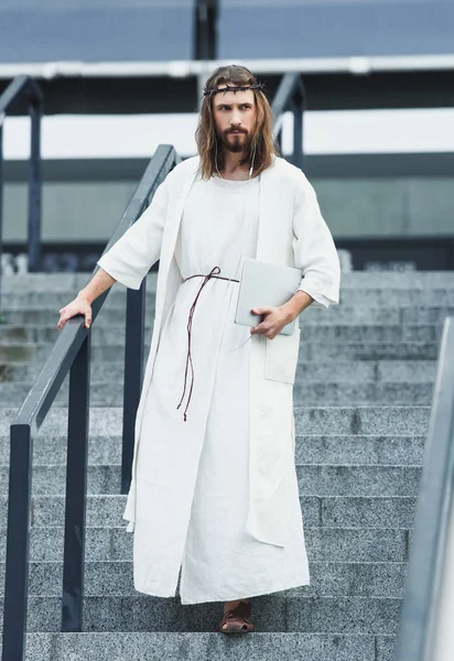 Серьезный Иисус в мантии и терновом венце, идущий по лестнице с ноутбуком и слушающий музыку в городе — стоковое фото
